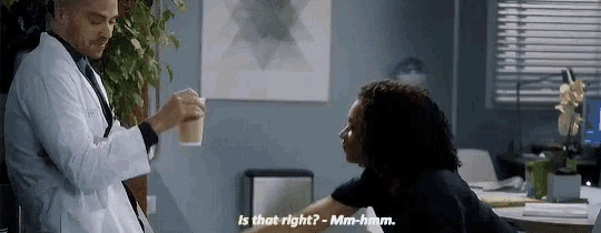 a man in a lab coat is holding a cup of coffee and talking to a woman in a black shirt .