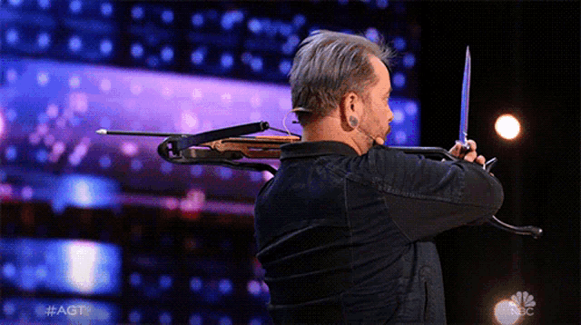a man is holding a crossbow and a knife in front of a nbc logo