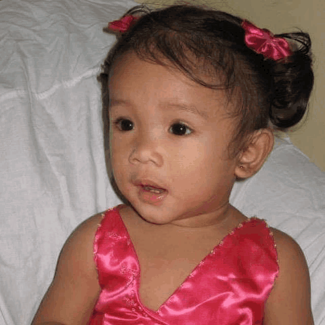 a little girl in a pink dress is sitting on a bed and looking at the camera .
