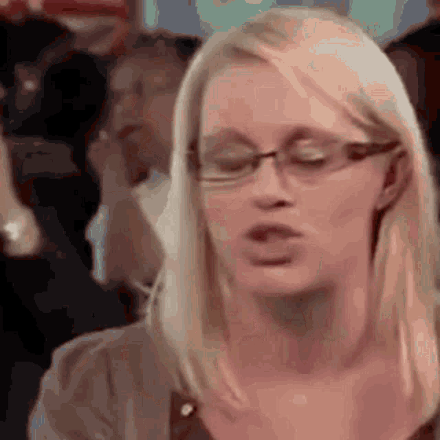 a woman wearing glasses is making a funny face while sitting in front of a crowd .