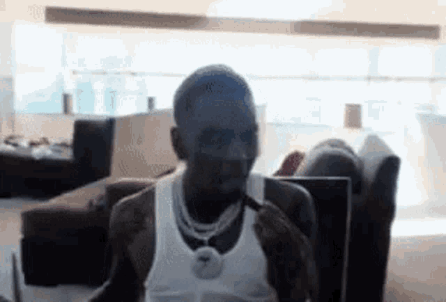 a man in a white tank top is sitting in a living room looking at a laptop .