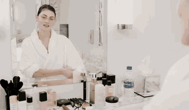 a woman in a bathrobe is looking at herself in a bathroom mirror with a bottle of aquafina water on the counter