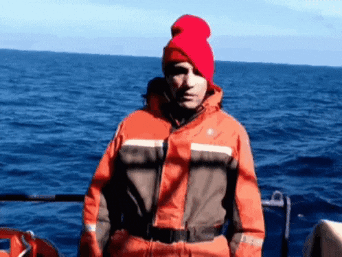 a man wearing an orange jacket and a red hat is standing on a boat in the ocean .