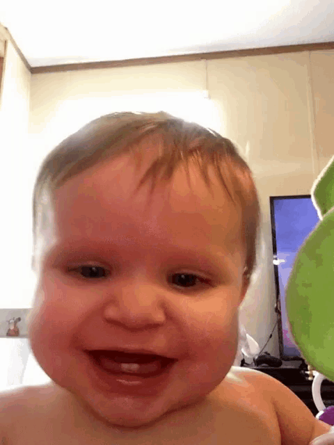 a baby is smiling and looking at the camera with a green stuffed animal in the background