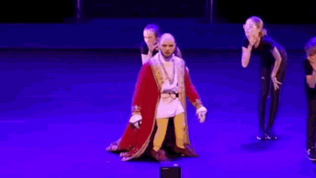 a man in a red robe is dancing on a stage with a group of people behind him .