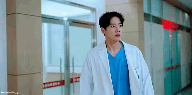 a man in a lab coat and blue scrub is walking down a hallway .