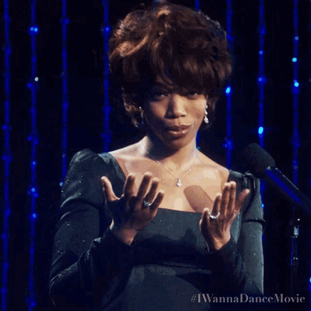 a woman in a black dress is standing in front of a microphone with her hands folded in front of her chest