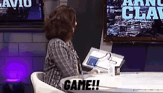 a woman is sitting at a table with a laptop in front of a television and says `` game '' .