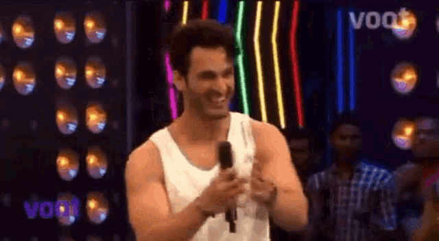 a man in a white tank top is holding a microphone and smiling while standing in front of a crowd .