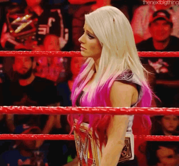 a woman with blonde hair and pink streaks is standing in a wrestling ring holding a championship belt .