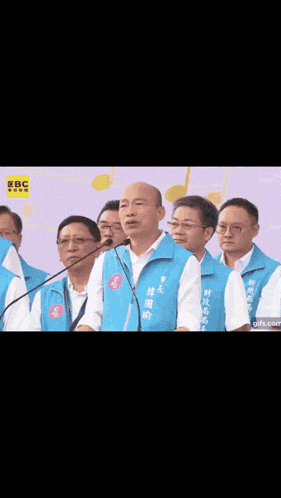a man in a blue vest is speaking into a microphone in front of a group of men