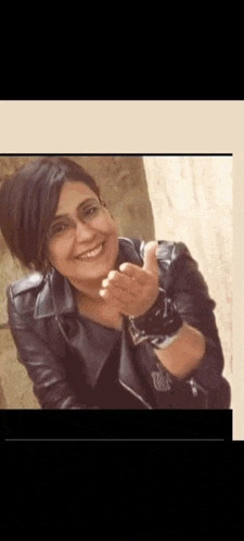 a woman wearing a leather jacket and glasses is smiling and giving a peace sign .