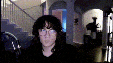 a woman wearing glasses stands in a dark room with stairs in the background