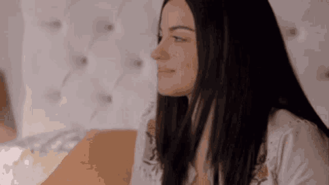 a woman with long black hair is sitting on a bed with a white headboard