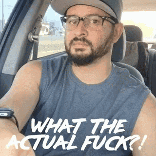 a man with a beard and glasses is sitting in a car wearing a shirt that says what the actual fuck ?