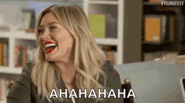 a woman is laughing while sitting in a chair in front of a bookshelf and says ahaha .