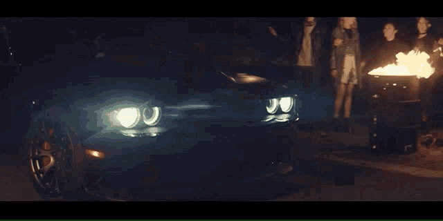 a group of people are standing around a fire pit while a car is parked in front of them