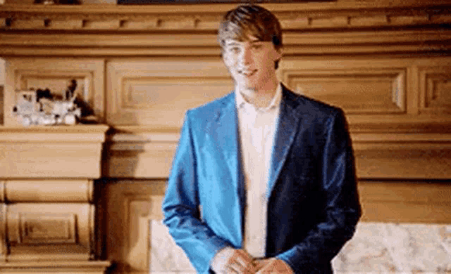 a young man in a blue jacket and white shirt is standing in front of a fireplace .