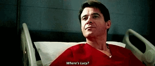 a man is laying in a hospital bed with a red shirt on and asking where 's lucy .