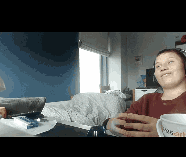 a person wearing headphones sits at a table with a box of cigarettes and a mug that says " was "