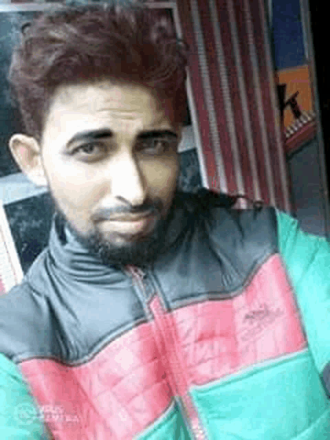 a man with a beard is wearing a red and green jacket and taking a selfie .
