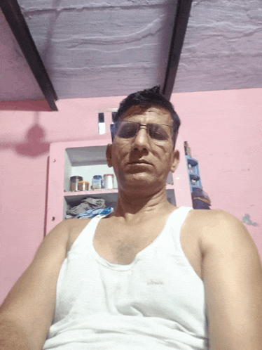 a man wearing glasses and a white tank top is sitting in a room