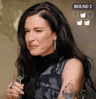 a woman with a tattoo on her arm is holding a shot glass in front of a round 2 sign