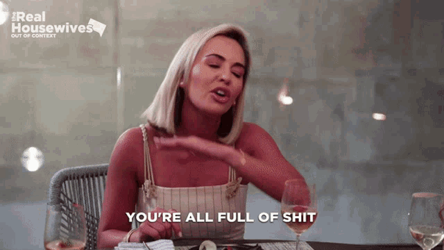a woman is sitting at a table with a glass of wine and says " you 're all full of shit "