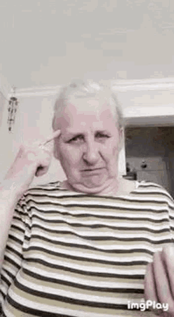 an elderly woman in a striped shirt is making a funny face with her finger to her forehead .