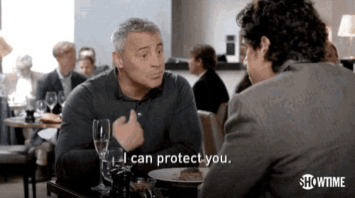 two men are sitting at a table in a restaurant talking to each other and one of them is saying `` i can protect you ''