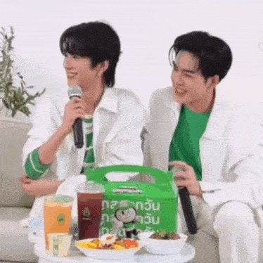 two men are sitting next to each other on a couch holding microphones and a bag of food .