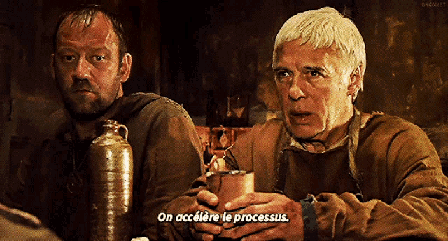 two men are sitting at a table and one of them is holding a cup that says on accelere le processus
