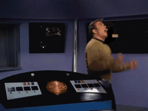 a man in a yellow sweater is standing in front of a purple wall and a control panel with a light on it