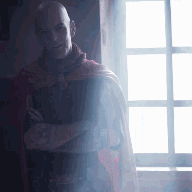 a bald man wearing a red cape stands in front of a window