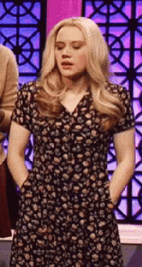 a woman in a floral dress is standing in front of a purple wall with her hands in her pockets .