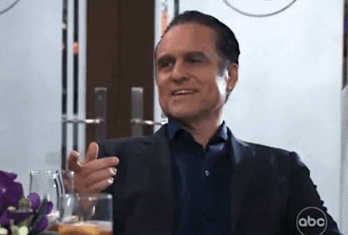 a man in a suit and blue shirt is sitting at a table with a glass of whiskey in his hand .