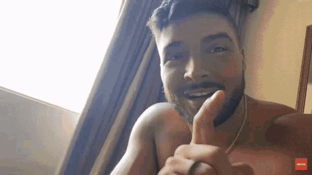 a shirtless man with a beard is giving a thumbs up in front of a window