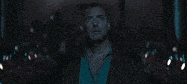a man in a blue shirt and brown jacket stands in a dark room