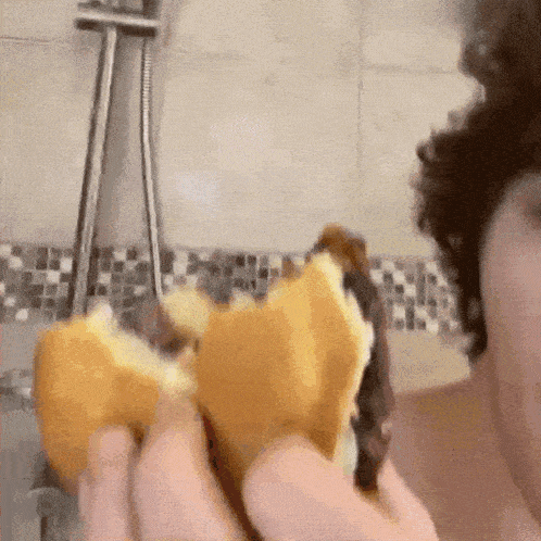a person is eating a sandwich in a shower with a shower head in the background