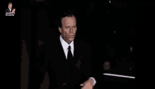 a man in a suit and tie is standing in a dark room with his hands on his chest .