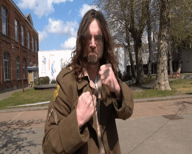 a man with long hair and glasses stands in front of a building
