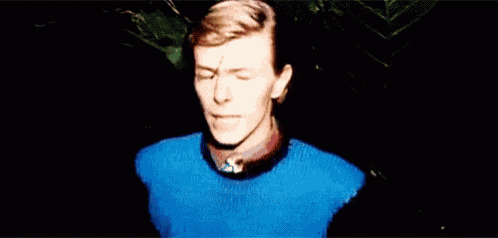 a man wearing a blue sweater and a red shirt is standing in a dark room with his eyes closed .