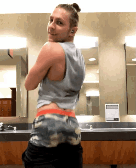 a man in a tank top and camo shorts is standing in front of a bathroom mirror