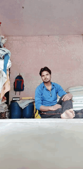 a man in a blue shirt sits on a bed with his feet crossed