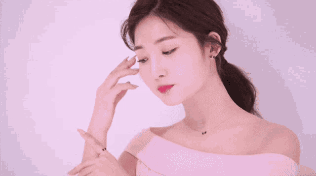 a woman wearing a pink off the shoulder top is touching her face with her hand