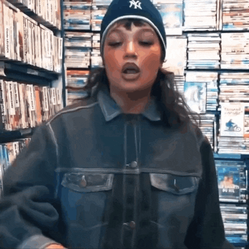 a woman wearing a ny hat and a denim jacket is standing in front of a stack of dvds