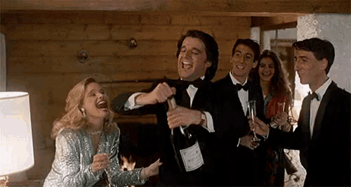 a man in a tuxedo is opening a bottle of champagne while a woman in a silver dress laughs .