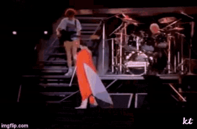 a man is playing a guitar on a stage with a drum set behind him .