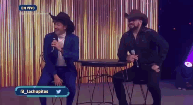two men wearing cowboy hats are sitting at a table holding microphones .