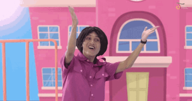a man in a purple shirt is standing in front of a pink building with his arms in the air